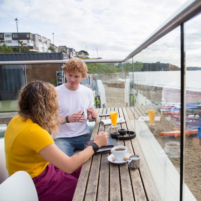 Falmouth University students sat on the terrace at Gylly Beach Cafe with the sea in the background